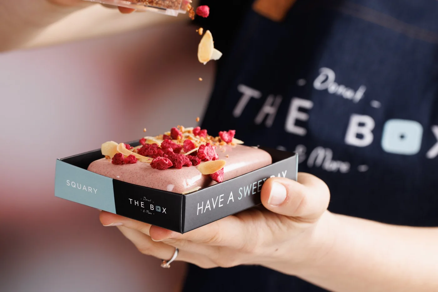 Hands sprinkling freeze-dried raspberries over a pink glazed square donut in a THE BOX gift box