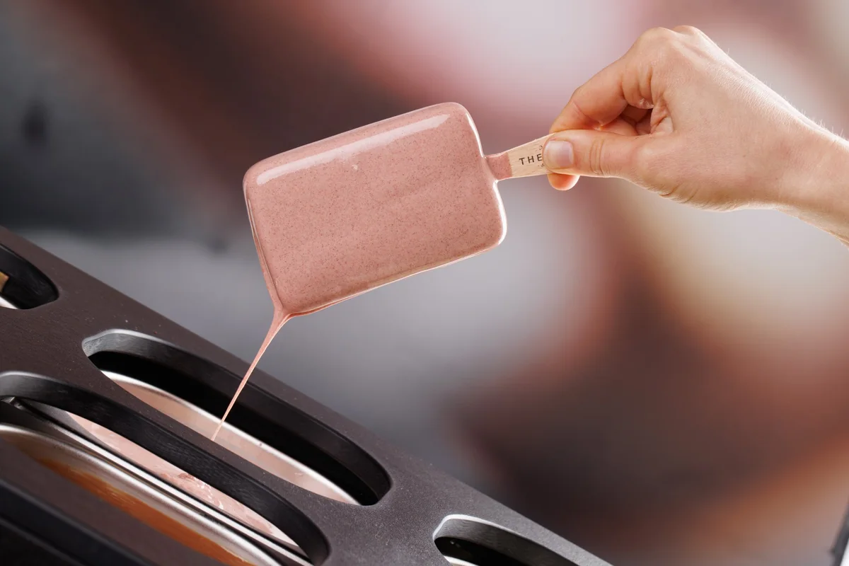 A square donut being dipped into pink glaze over the glazing machine